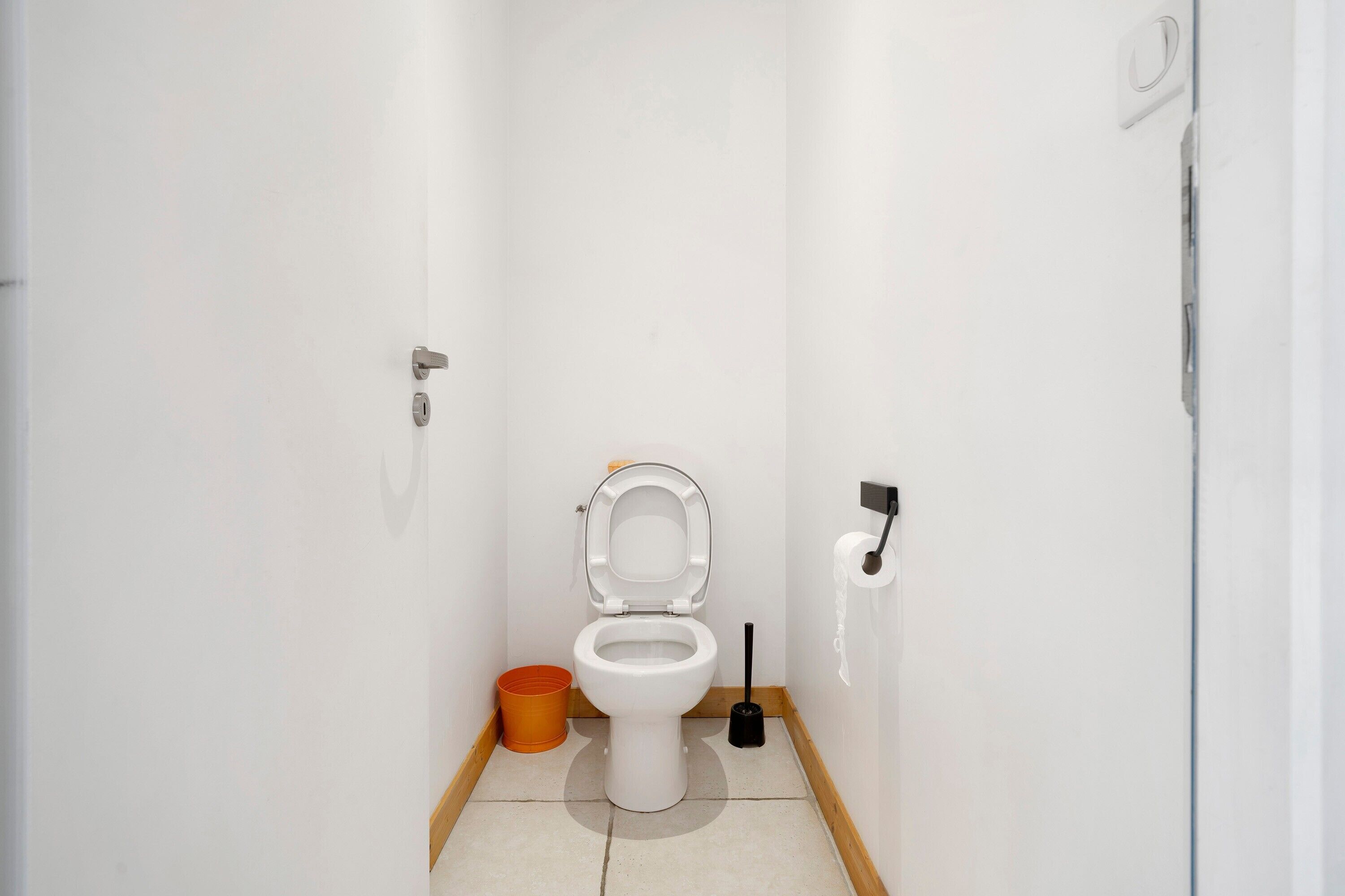 A small, white bathroom with a toilet, a trash bin, and a toilet brush.