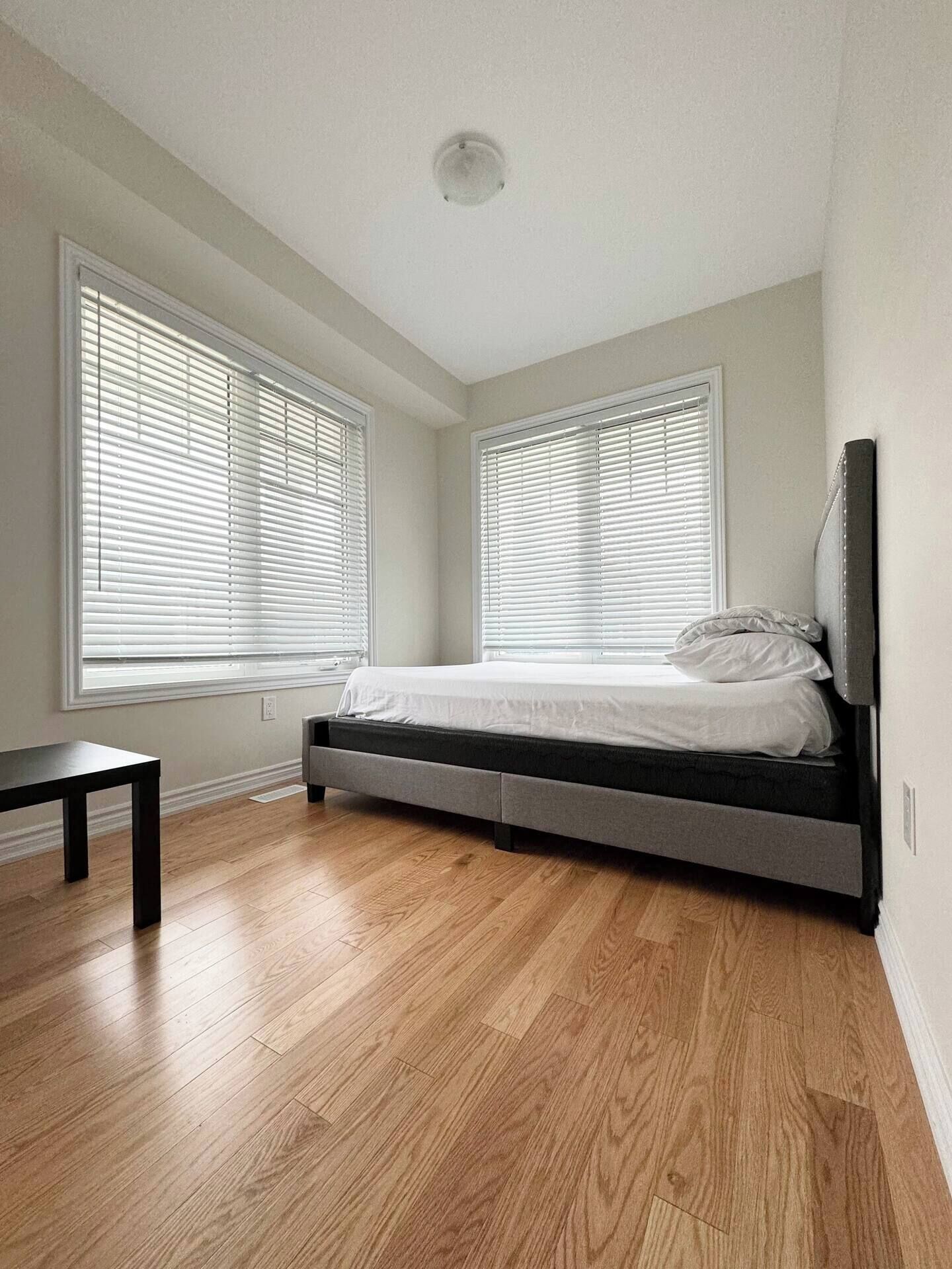 A bedroom with a bed, a bedside table, and two windows with blinds.
