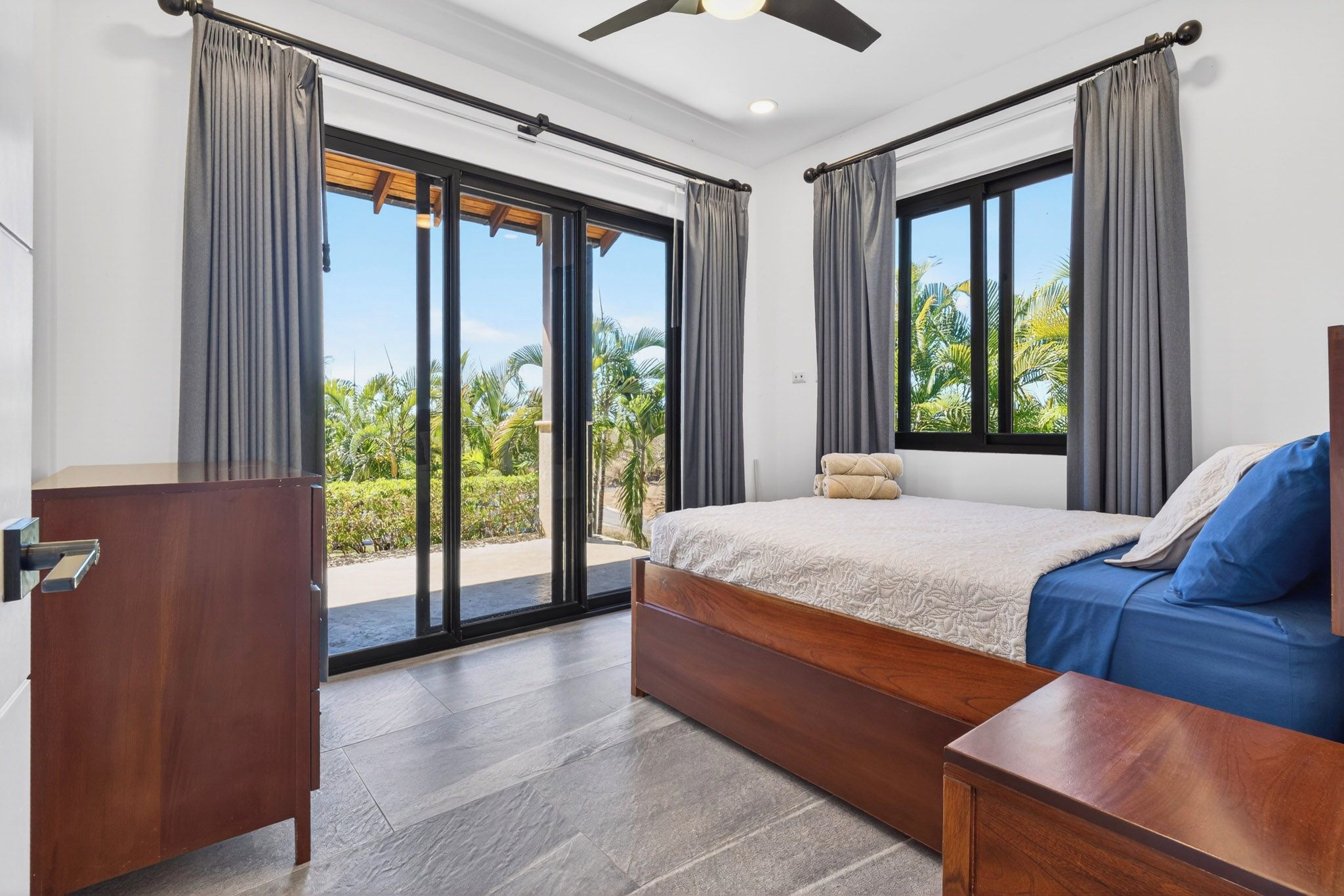 Ein Schlafzimmer mit einem Bett, einem Schreibtisch und Blick auf Palmen durch große Fenster.