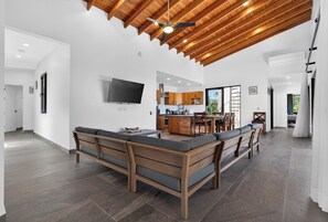 A spacious living area with a wooden ceiling, a large sofa, a kitchen with wooden cabinets, and a TV mounted on the wall.