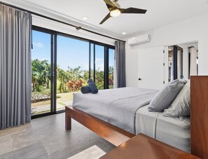 A bedroom with a bed, a bedside table, a ceiling fan, and a view of a garden through large glass doors.