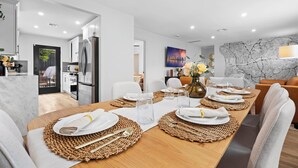 A modern dining area with a long wooden table set for a meal, white chairs, and a cityscape painting on the wall. - Hot Tub/Pool/Fire Pit/10 Mins to Beach (San Diego)
