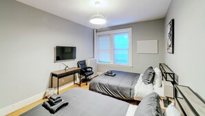 Ein modernes Schlafzimmer mit zwei Betten, einem Schreibtisch, einem Sessel, einem Fernseher und einem Fenster mit Jalousien.
