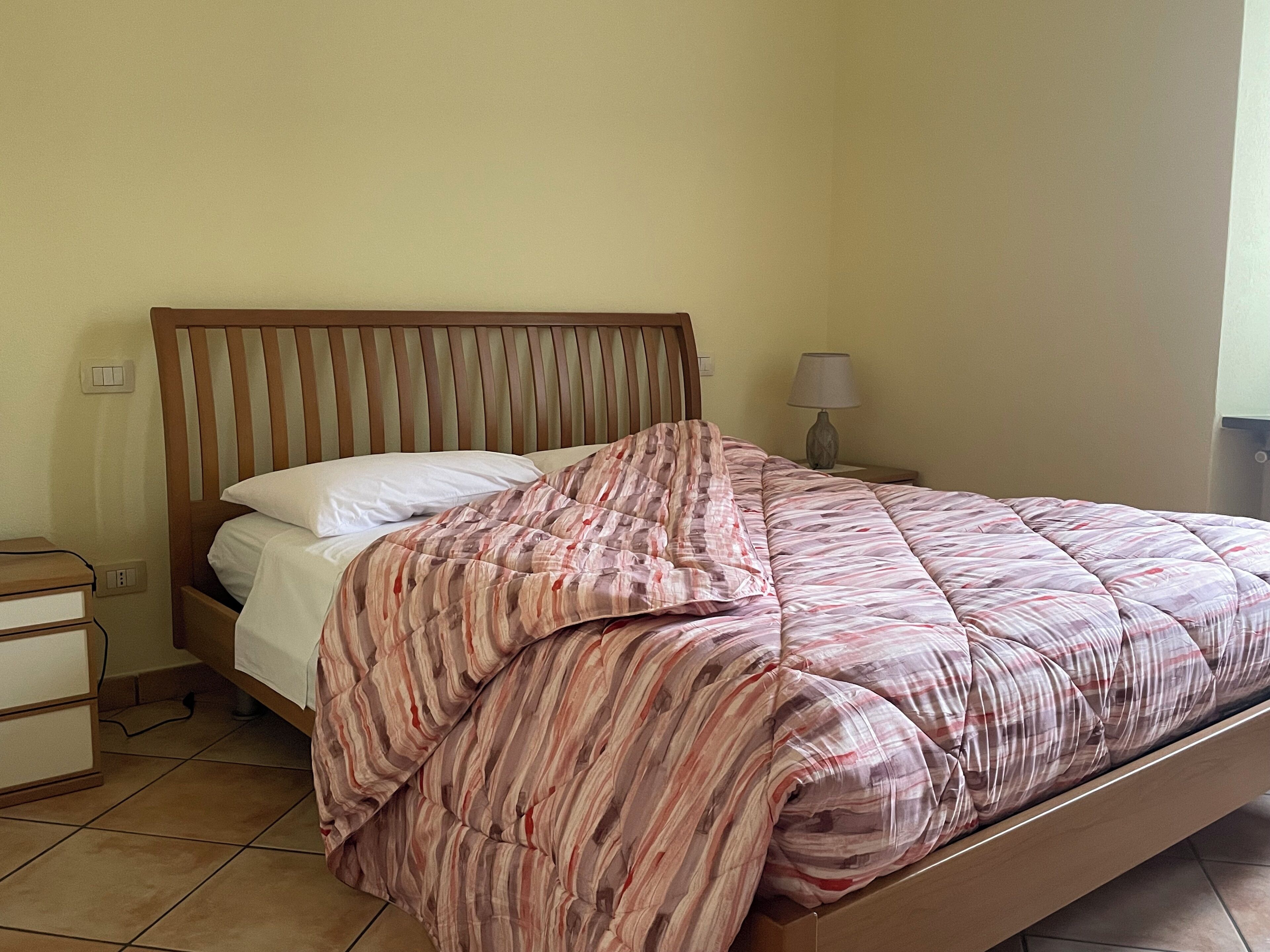 Een eenpersoonsbed met een gestreept sprei, een houten hoofdbord, een nachtkastje met een lamp en een nachttafeltje met een lamp.