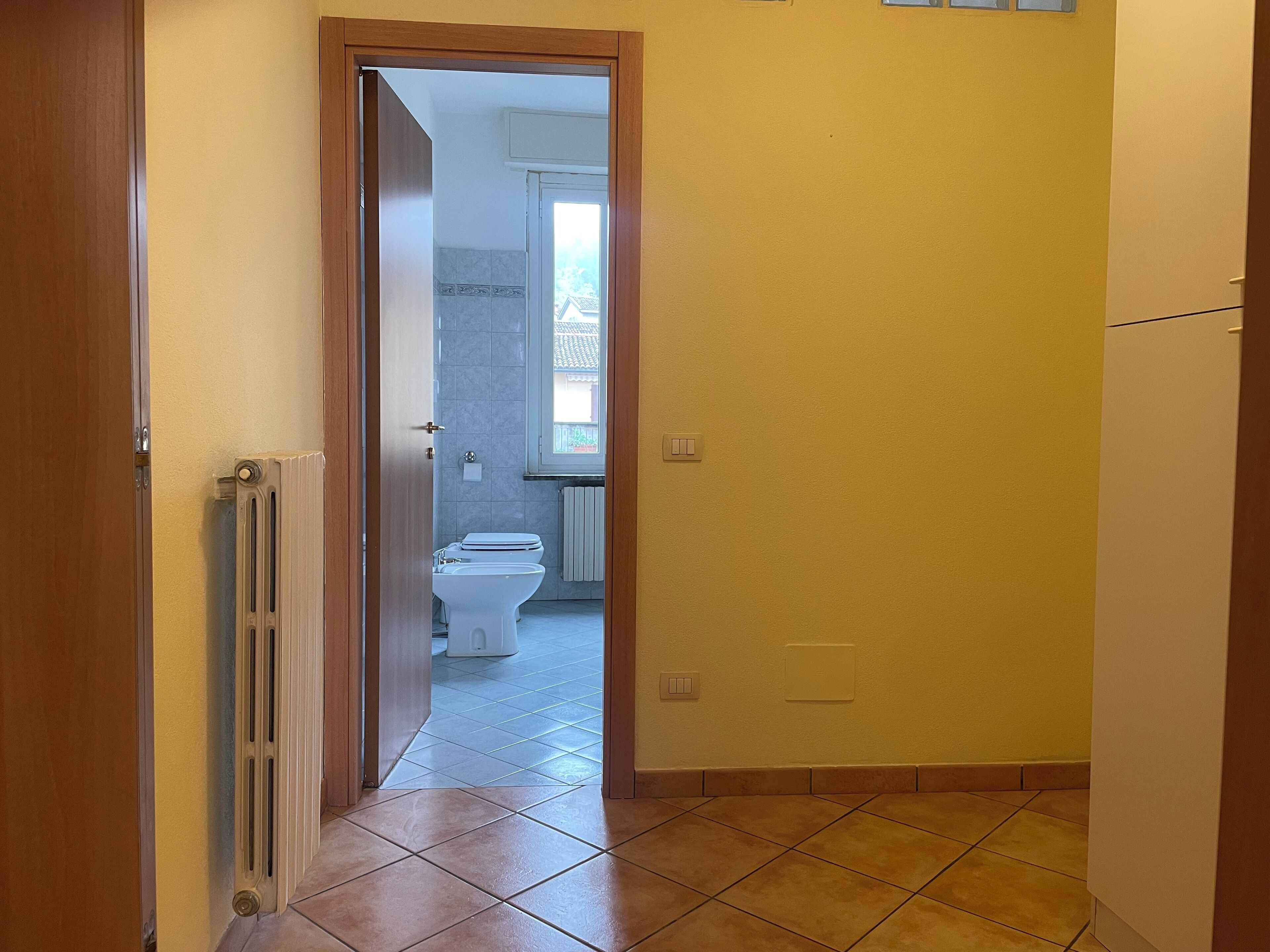 Een smalle gang met een deur die leidt naar een badkamer met toilet en radiator.