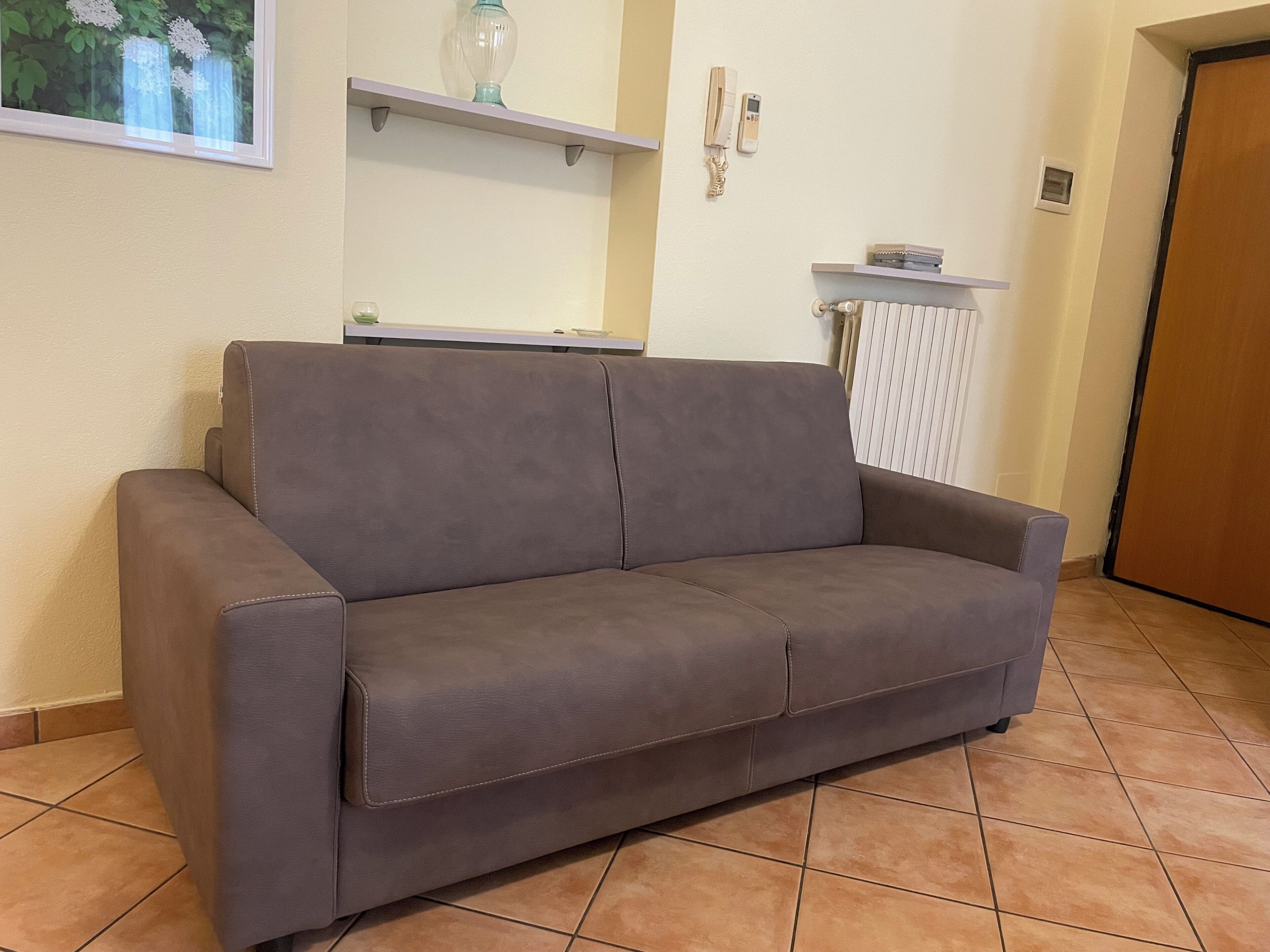 Een bruine bank in een kamer met betegelde vloer, een witte radiator en een deur.