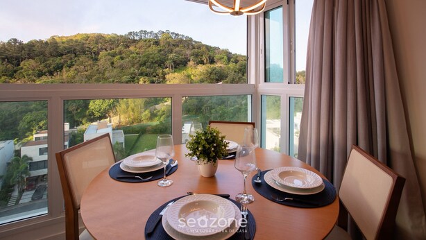 A dining table set with plates, glasses, and a vase of flowers, overlooking a lush hillside. - Modern and Complete Apartments in Itajai - RVC (Itajai)