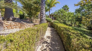 A pathway flanked by neatly trimmed hedges and palm trees, leading to a stone wall and a building with a tiled roof. - Villa Caripira by Villa Plus (Albufeira)