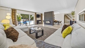 A modern living room with a sofa, coffee table, and a fireplace. There is a pool visible through the glass doors. - Villa Caripira by Villa Plus (Albufeira)