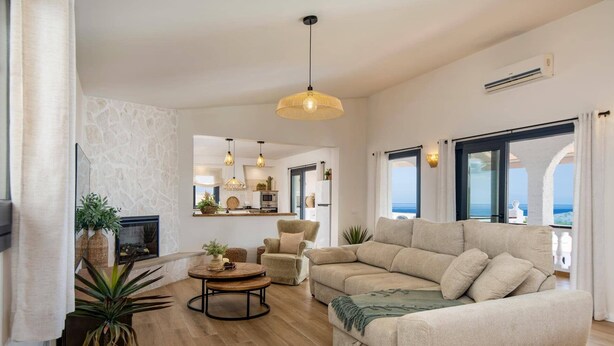 Een ruime woonkamer met een grote beige bank, een ronde salontafel en een open haard. Er staan potplanten en er is uitzicht op de oceaan door grote ramen.