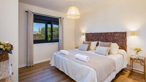 Een slaapkamer met een groot bed, een raam met gordijnen, een houten hoofdbord en een nachtkastje met een bloemenvaas.