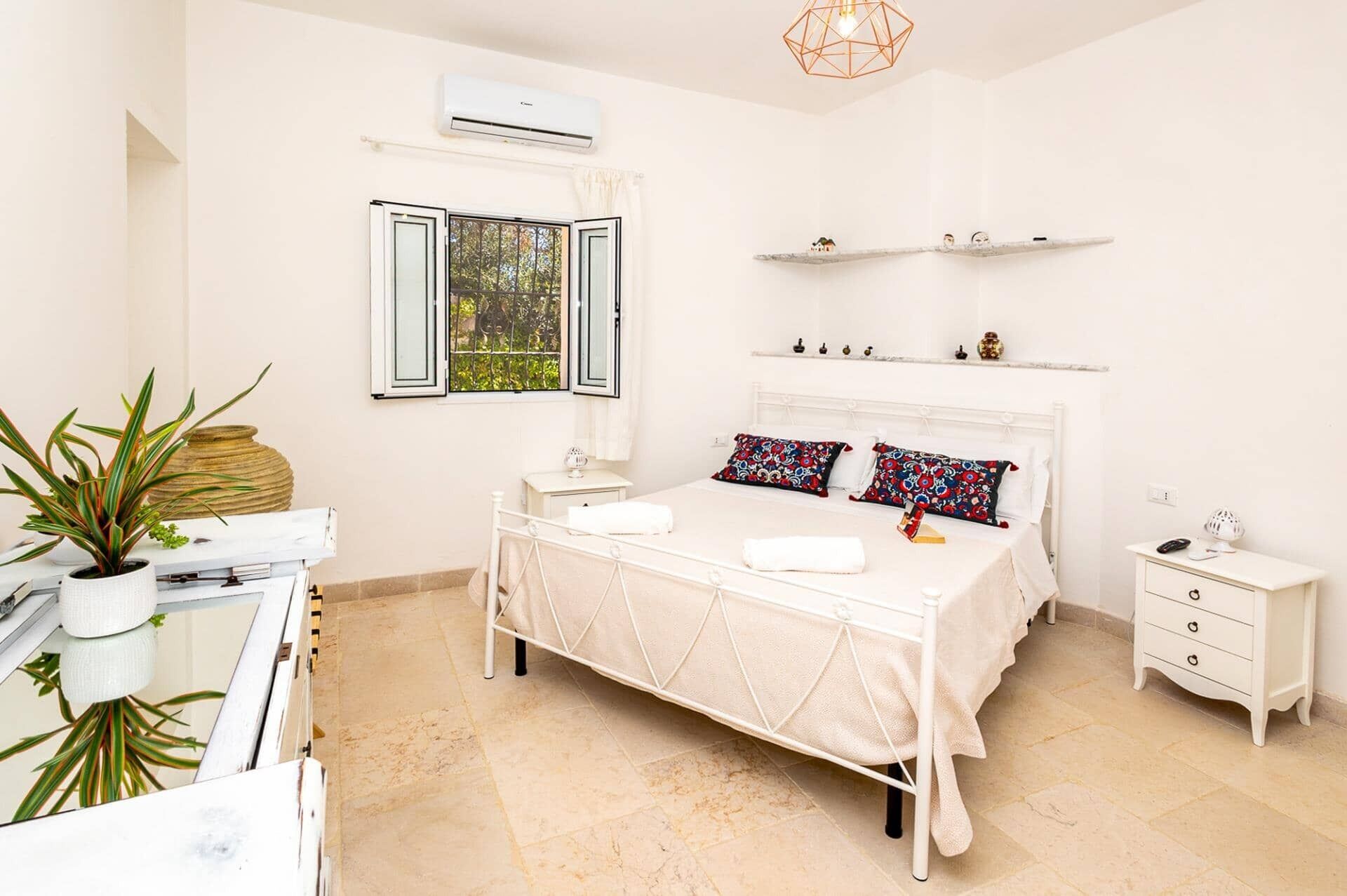A bedroom with a bed, bedside tables, a desk, and a chair. There is a window with curtains, a shelf with decorative items, and an air conditioning unit.