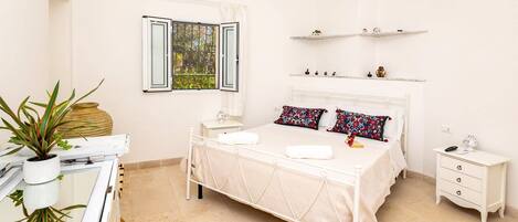 A bedroom with a bed, bedside tables, a desk, and a chair. There is a window with curtains, a shelf with decorative items, and an air conditioning unit.