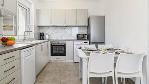Een compacte keuken met witte kasten, een ingebouwde oven, magnetron en vaatwasser. Er staat een eettafel met stoelen en een fruitschaal op het aanrecht.