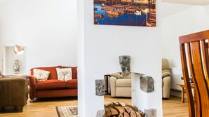 Un salon avec un canapé rouge, un fauteuil beige et une chaise en bois. On y trouve une cheminée avec un manteau en pierre et un feu de bois. Un tableau représentant un port est accroché au mur.