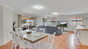 Un vaste espace de vie comprenant un coin repas avec une table, des chaises blanches, un canapé gris, un téléviseur et un coin cuisine avec un bar.