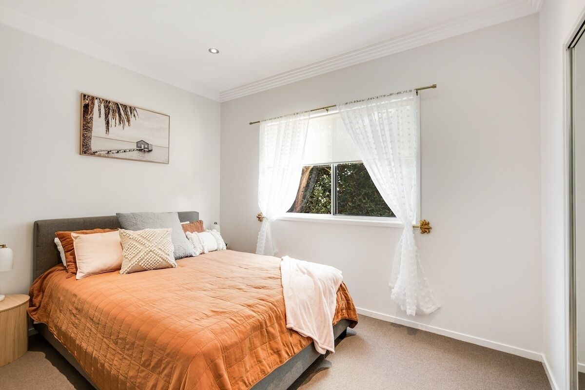 Een slaapkamer met een bed, een raam met dunne gordijnen, een nachtkastje en een aan de muur hangend schilderij.