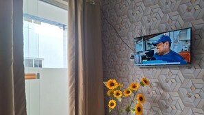 A room with a TV mounted on a textured wall, a window with curtains, and a vase with sunflowers. - Apartamento novo em João Pessoa  (João Pessoa)