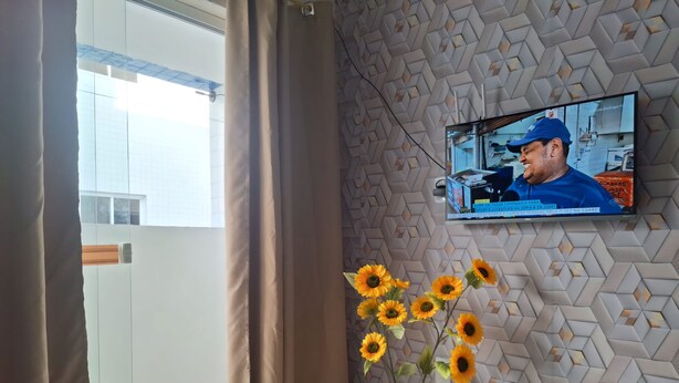 A room with a TV mounted on a textured wall, a window with curtains, and a vase with sunflowers. - Apartamento novo em João Pessoa  (João Pessoa)