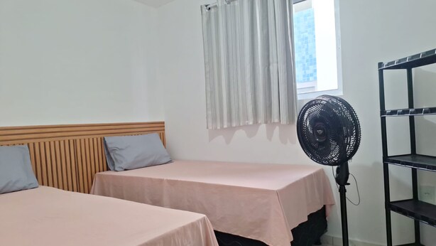 A room with two beds, a window with curtains, a standing fan, and a black shelf unit. - Apartamento novo em João Pessoa  (João Pessoa)