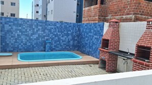 A small outdoor area with a swimming pool, a brick barbecue grill, and a tiled wall. - Apartamento novo em João Pessoa  (João Pessoa)