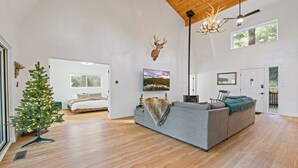 Un salon spacieux avec un grand canapé gris, un plafond en bois, une tête de cerf accrochée au mur, un sapin de Noël et un tableau représentant un paysage.