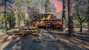 Une cabane dotée d’une terrasse, entourée de grands arbres et accessible par un chemin de gravier menant à une table de pique-nique.