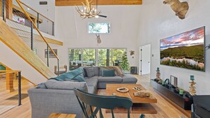 Un vaste espace de vie agrémenté d’un escalier en bois, d’un grand canapé d’angle gris, d’une table à manger et d’une tête de cerf fixée au mur.