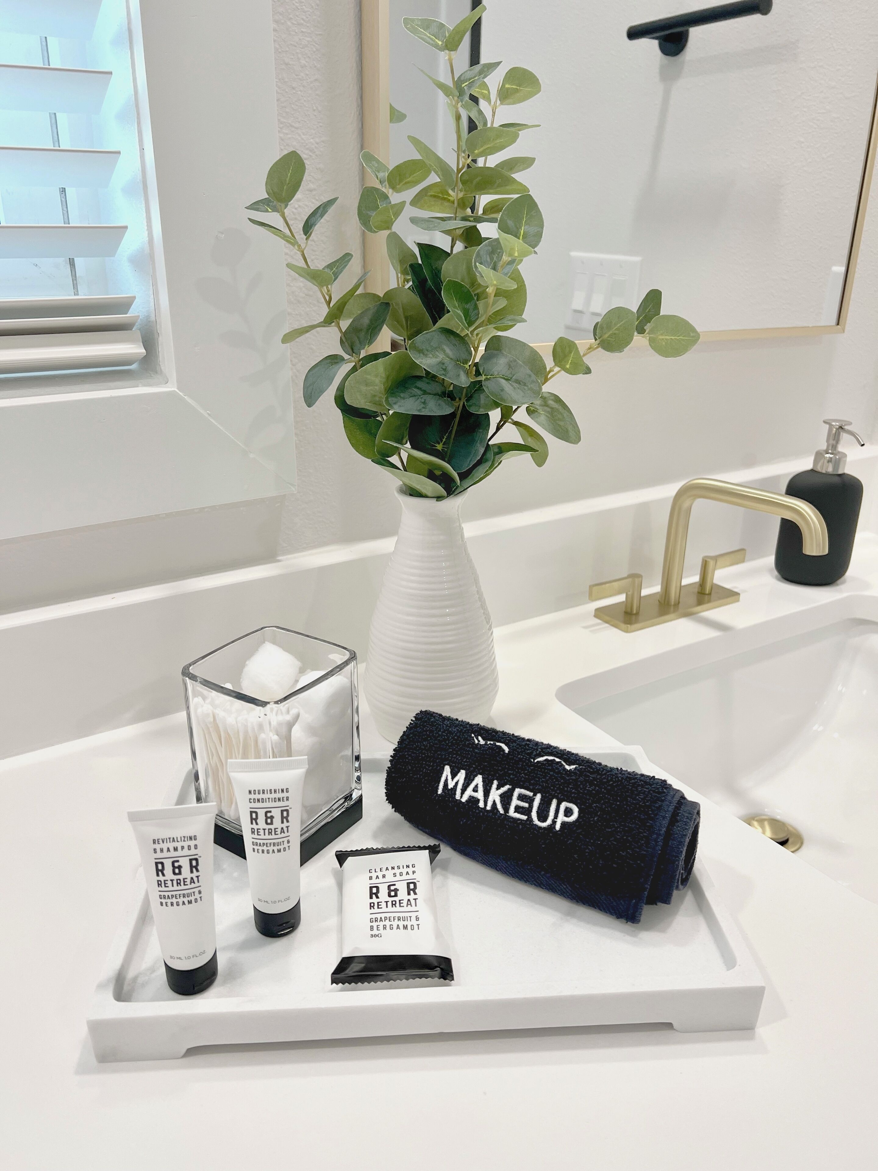 Un baño con lavamanos blanco, un espejo, una planta en un florero blanco y una bandeja con artículos de aseo que incluye una toalla de maquillaje, champú y jabón.