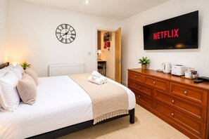 Et hotelværelse med en seng, en trækommode, et vægur, et fjernsyn der viser 'NETFLIX', og en dør til et badeværelse.