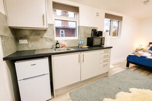 Eine kompakte Küche mit Kühlschrank, Spüle und Mikrowelle. Über der Spüle befindet sich ein Fenster, und im angrenzenden Raum ist ein Bett zu sehen.