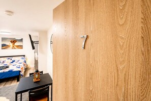 Ein Zimmer mit Holztür, einem Bett mit blumigem Überwurf, einem kleinen Tisch mit Lampe und einem Bild an der Wand.