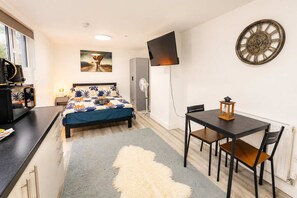 A compact room with a bed, a desk, a table with chairs, a TV mounted on the wall, and a decorative wall clock. - Trendy Roath Studio (Cardiff)