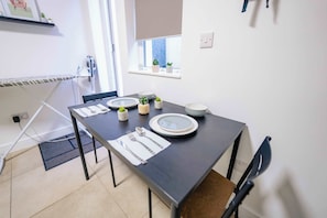 A dining area with a table set for two, featuring white plates, a fork, and a knife. There are small potted plants on the windowsill. - Modern Studio with Fast Wifi! (Cardiff)