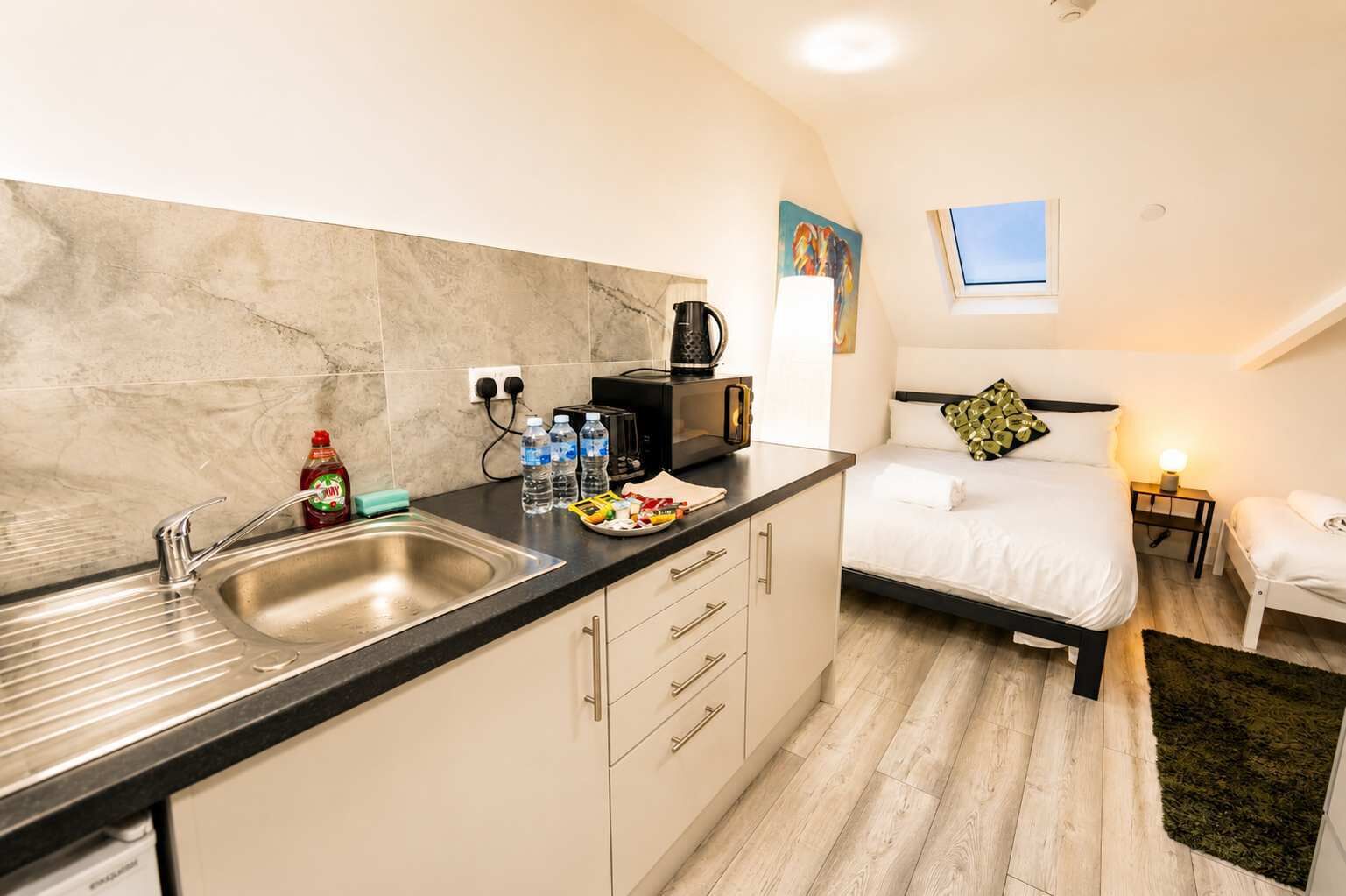 A compact hotel room with a kitchenette, a bed, a bedside table, and a small window.