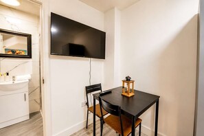Een kleine kamer met een flatscreen televisie aan de muur, een houten tafel met een lantaarn, een stoel en een wit wastafelmeubel met spiegel.
