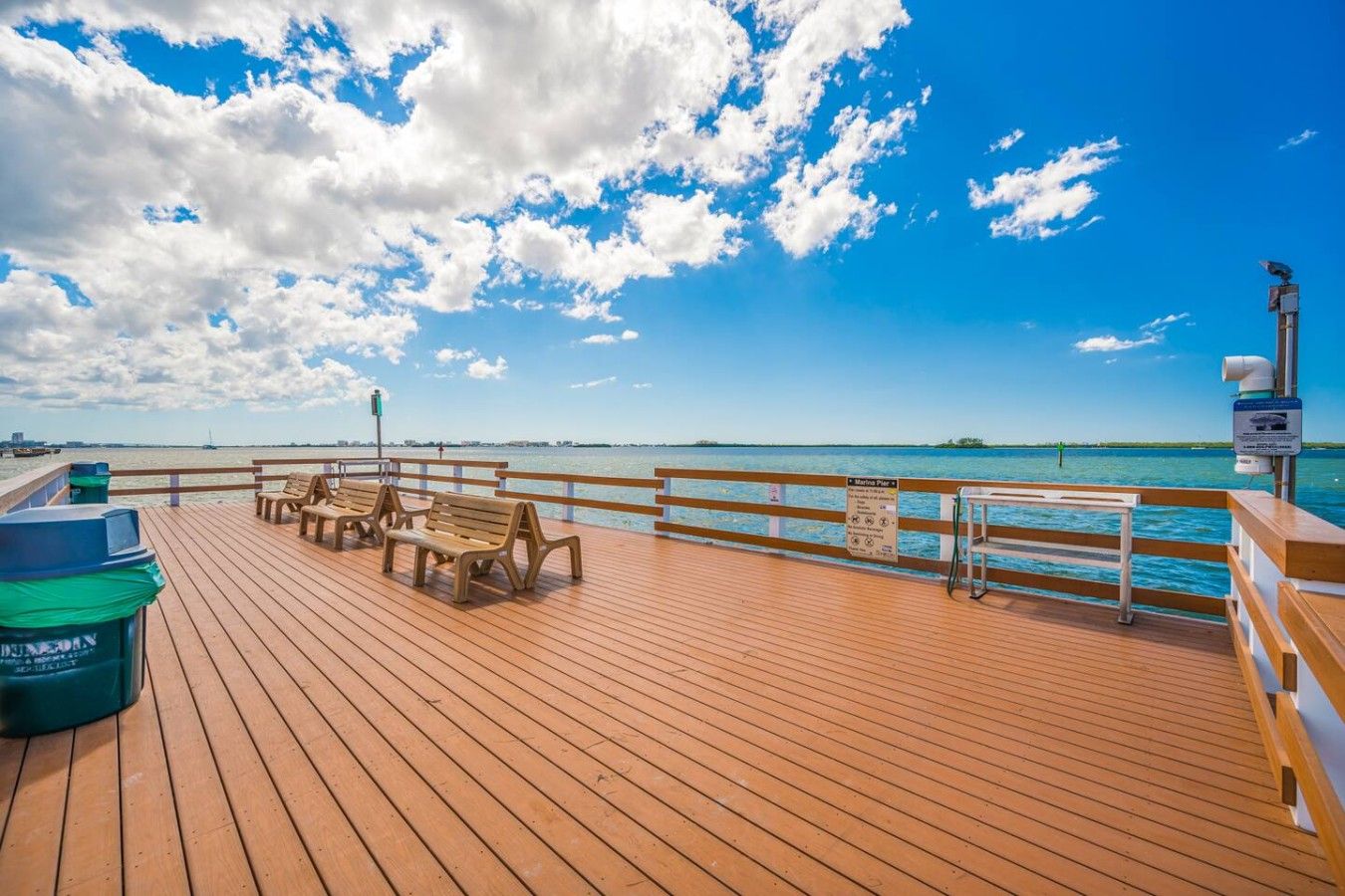 Eine Holzterrasse mit Bänken und einem Mülleimer, mit Blick auf ein Gewässer unter blauem Himmel.
