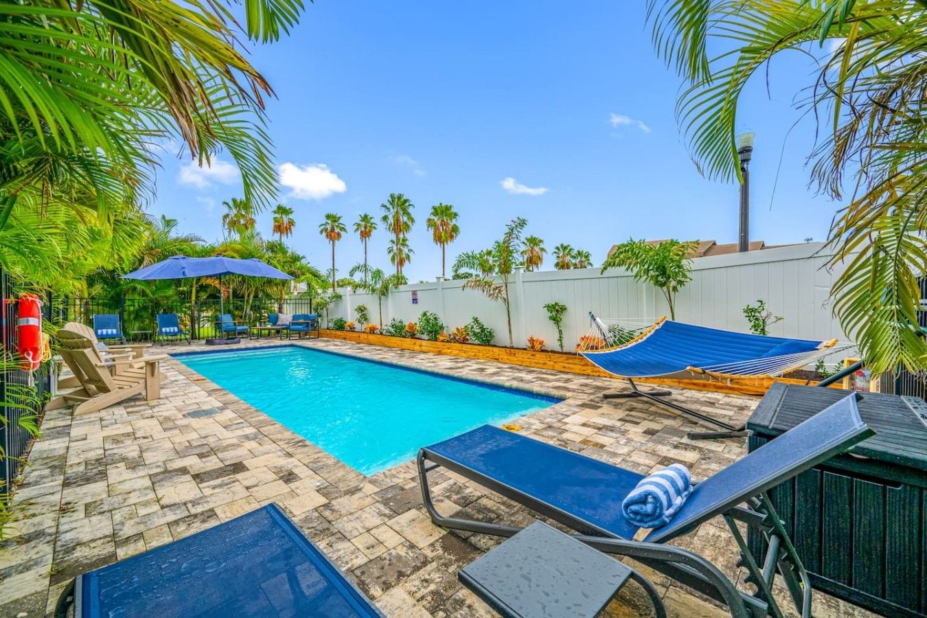 A swimming pool with lounge chairs and a hammock, surrounded by palm trees and a white wall.