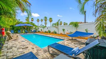 A swimming pool with lounge chairs and a hammock, surrounded by palm trees and a white wall.