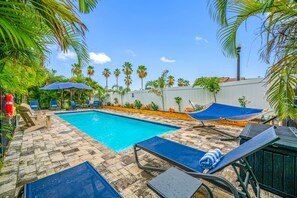 Piscina com cadeiras de descanso e rede, cercada por palmeiras e uma parede branca.