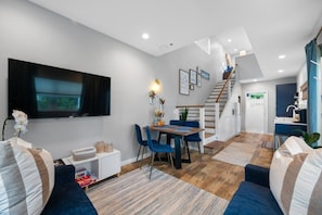 A modern living room with a flat-screen TV, a dining area with blue chairs, a staircase, and a kitchen area. - Walk Downtown | Private Patio | Pool | Pets | Skye (Dunedin)
