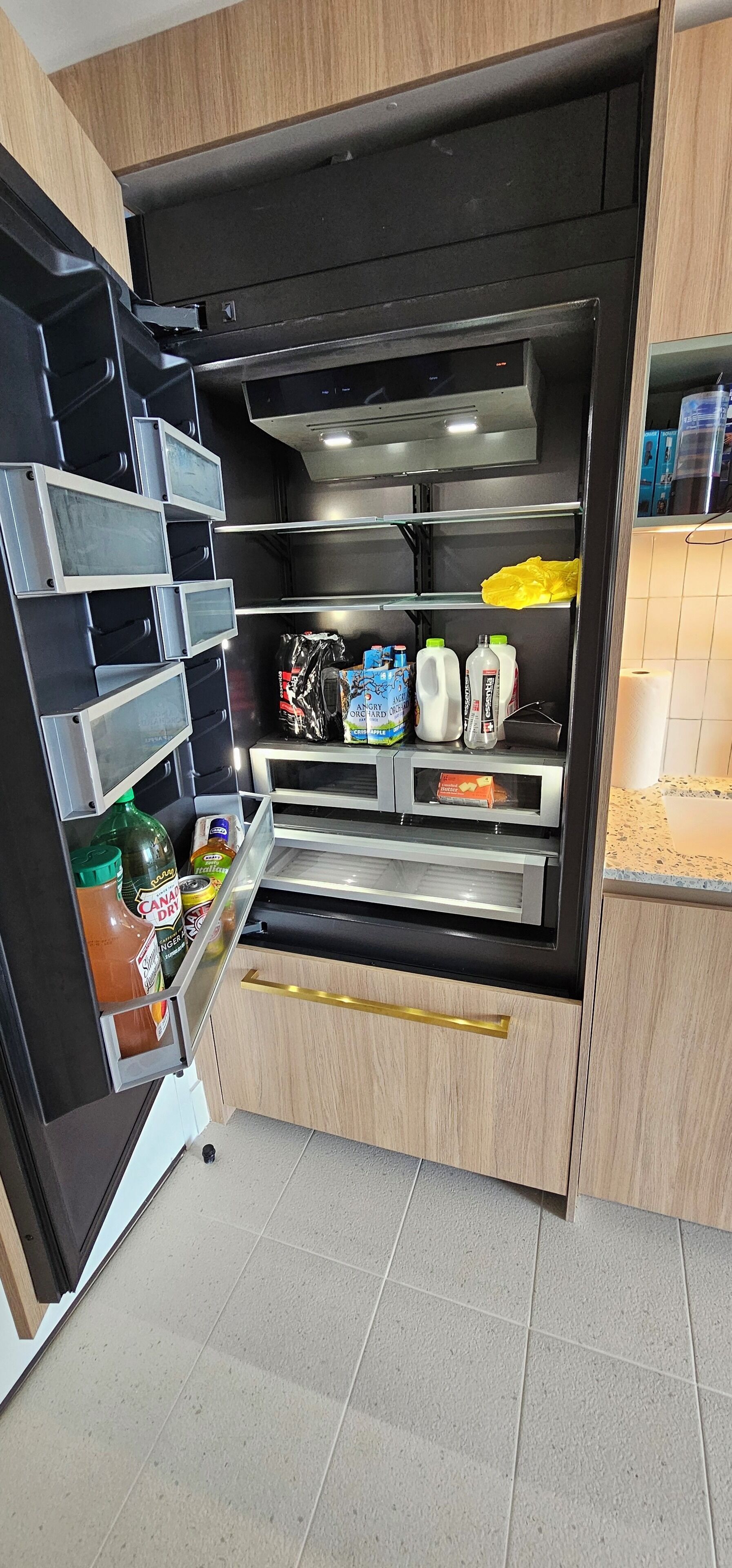 A kitchen with a built-in refrigerator, a microwave, and a shelf with various food items.
