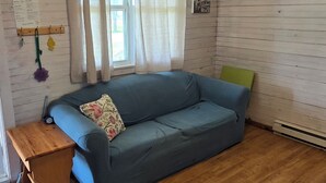 A room with a blue sofa, wooden beams on the ceiling, a window with curtains, a small table, and a wall sign. - Cabin - Older/Simple/Rustic/No Frills/Budget 2 Bedroom - Near Cabot Beach Park (Kensington)