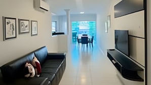 Ein modernes Wohnzimmer mit einer schwarzen Couch, einem flachbildfernseher an der Wand und einem Essbereich im Hintergrund.