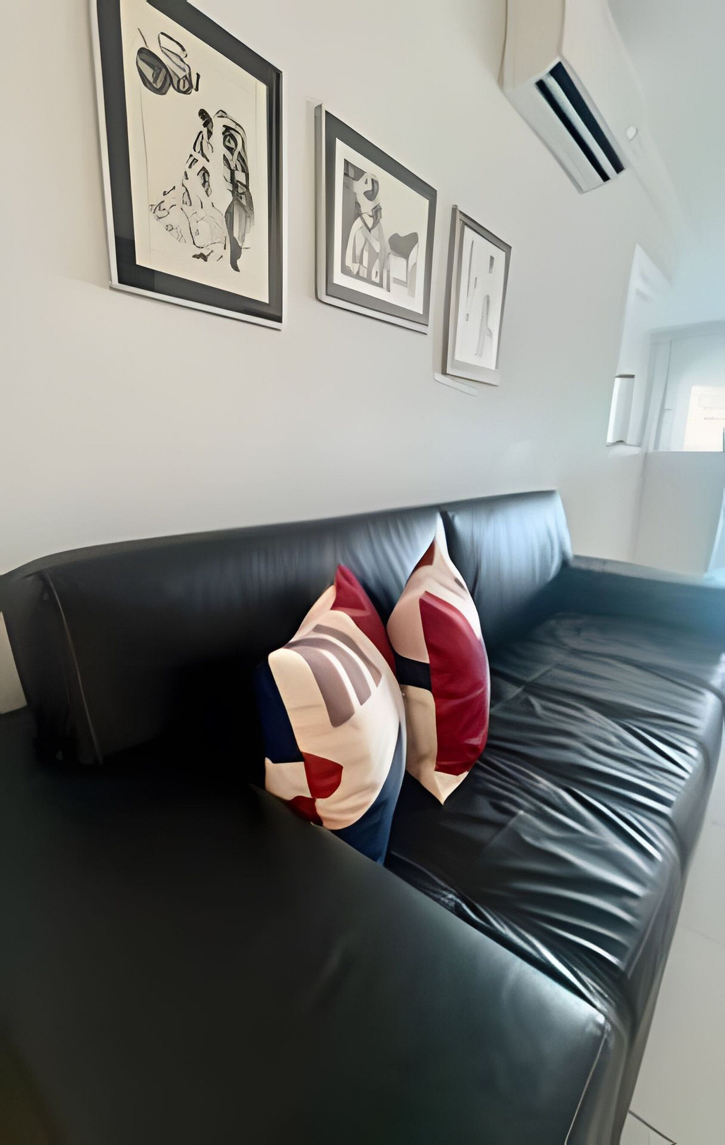 A black leather sofa with colorful geometric-patterned pillows, framed artwork on the wall, and a window with natural light.