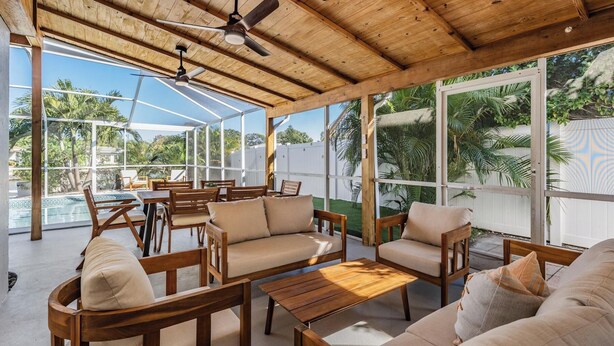 An outdoor patio with wooden furniture, a ceiling fan, and large windows offering views of palm trees and a pool. - Modern Waterfront Home w/ Pool Near Airport & South Tampa Hotspots (Tampa)