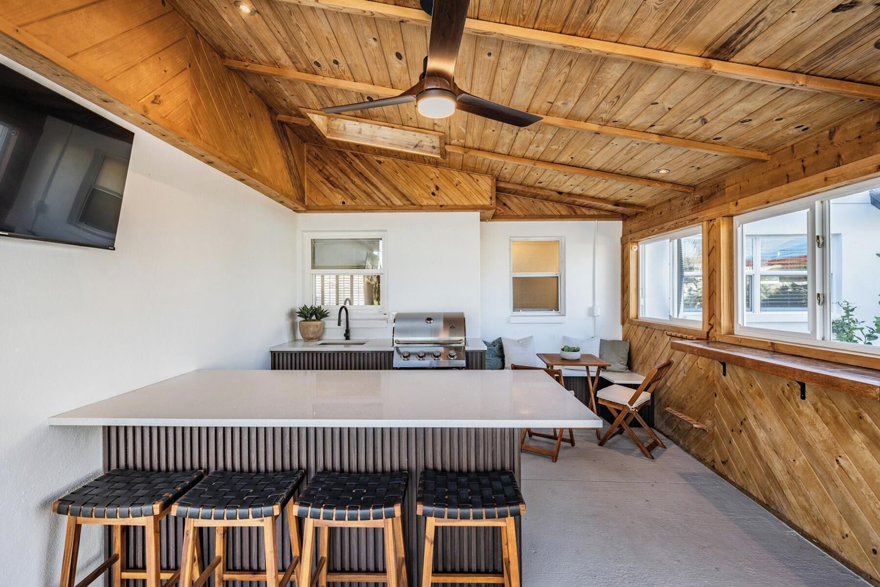 A modern kitchen with a wooden ceiling, a central island with bar stools, and large windows providing natural light.