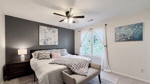 A bedroom with a bed, a nightstand, a ceiling fan, a window with curtains, and two wall-mounted paintings. - Perfect Location, Minutes to everything Alpharetta & Johns Creek (Alpharetta)
