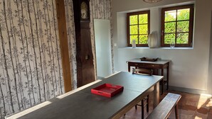 A dining area with a long table, benches, a clock, and two windows with greenery outside.