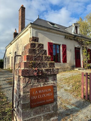 A rustic building with a brick staircase, red shutters, and a sign reading "LA MAISON DU CLOCHER".